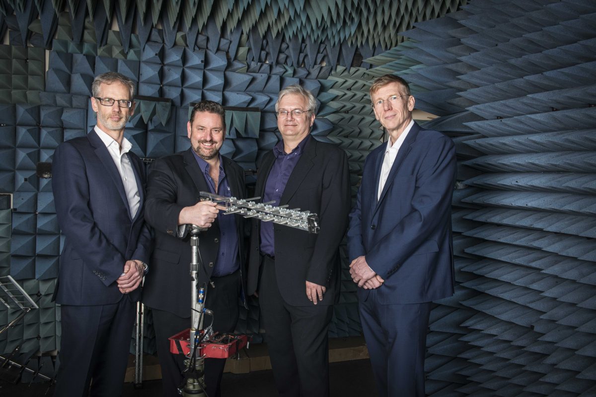 Four men in suits stood in a room with spikes on the wall showing some TV aerial equipment