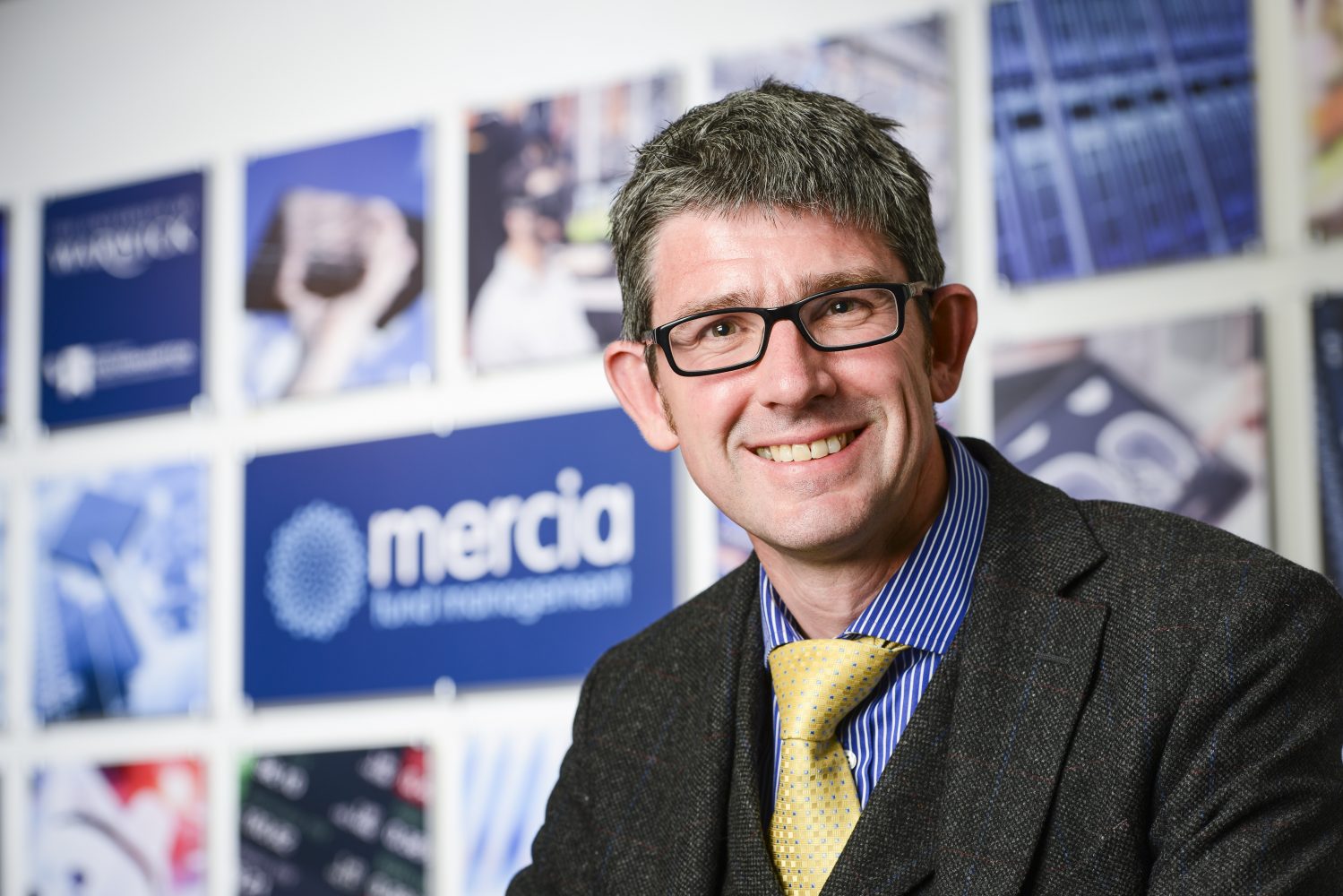 Man named Mark Payton with glasses and black jacket, yellow tie sitting in front of posters stating Mercia.