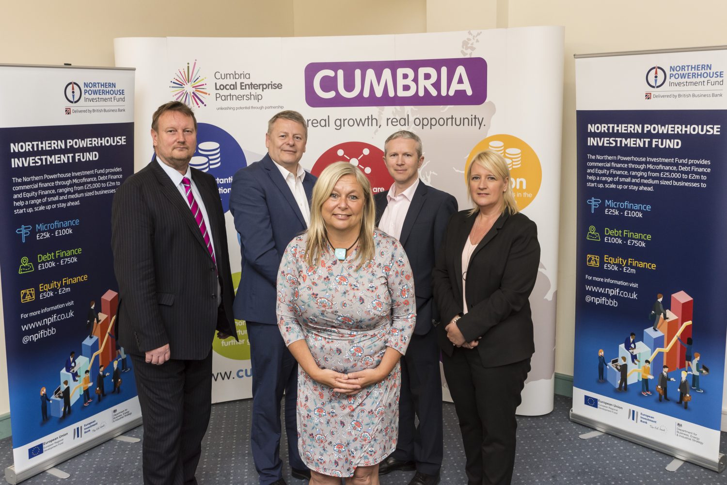 Five people standing behind Northern Powerhouse point of sale. There are two women in the group. The woman in the front has long blond hair and is wearing a grey patterned dress. The woman in the group to the far left is wearing a black trouser suit. There is a man to the far left wearing a red stripey tie and black suit.