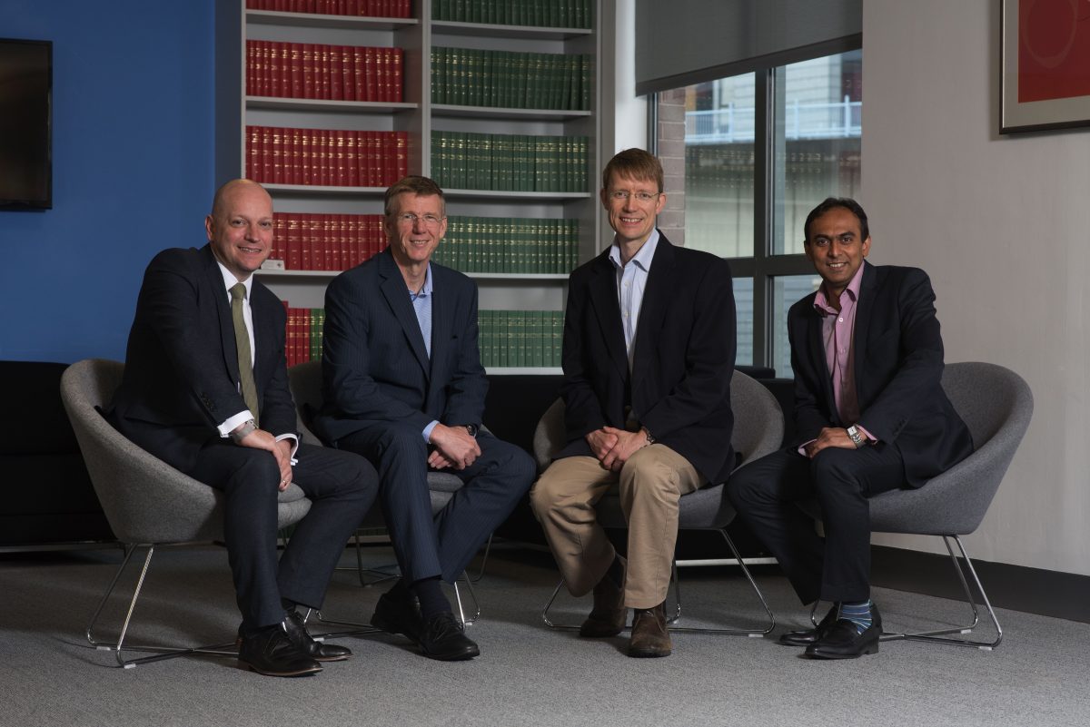 Four men sitting in grey chairs in an office in front of bookshelves.