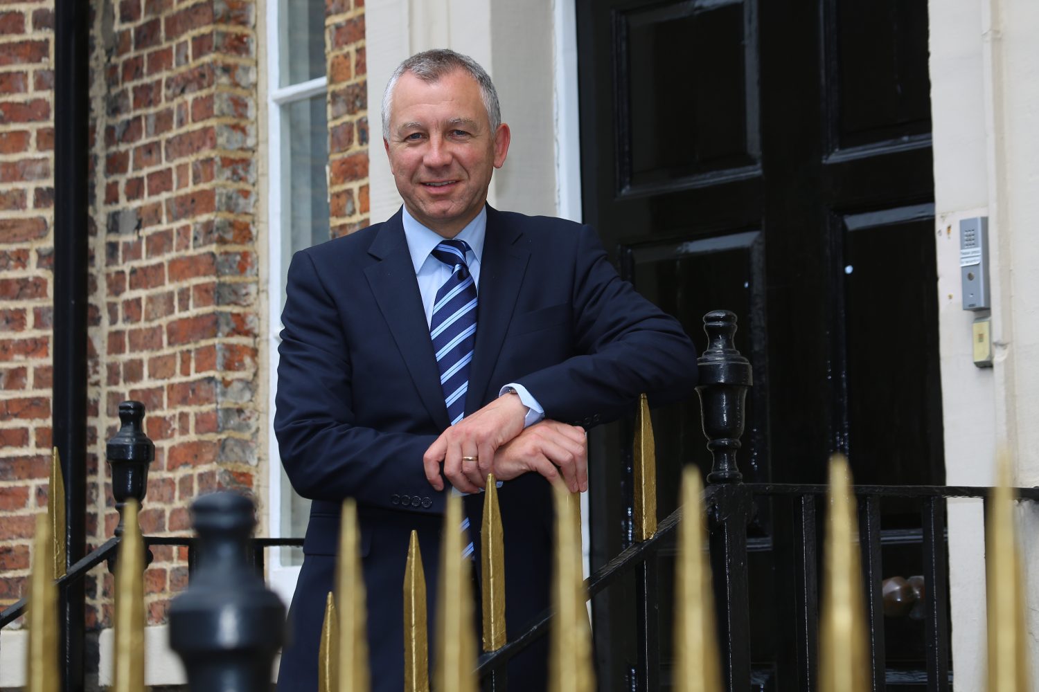 Head and chest shot of Keith Barnes standing in front of black painted door. He is holding onto some gold coloured railings.