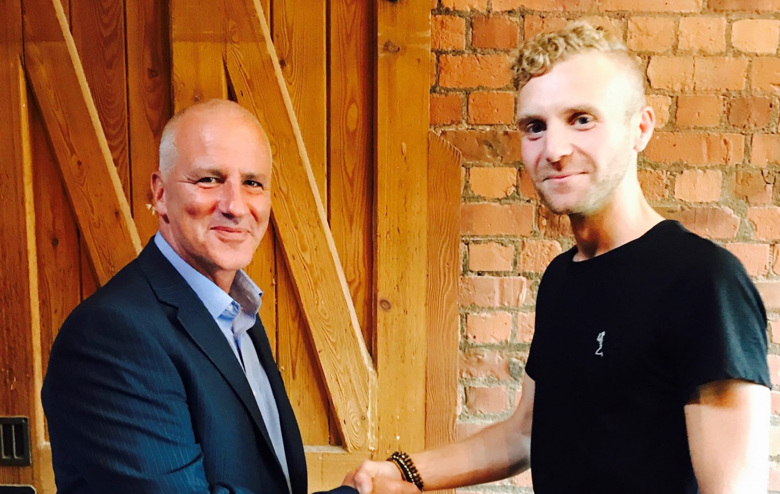 Two men shaking hands in a room with red brick wall in the background. One man is wearing a black tee shirt and the other man is wearing a blue suit.