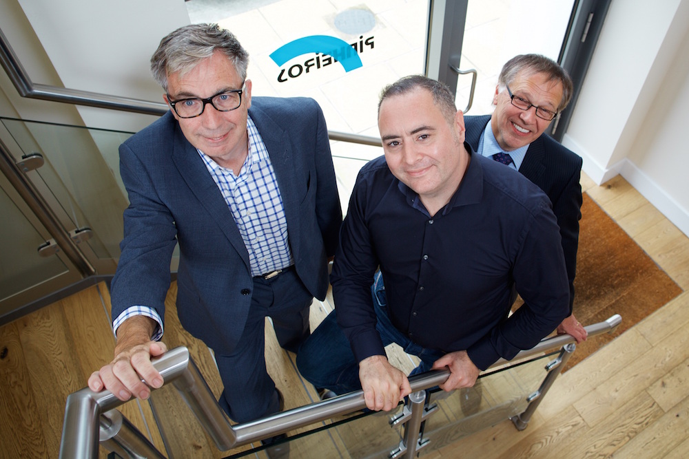 Three men standing on internal stairs. All the men have their hands on the handrail. Two men are wearing glasses and suits.