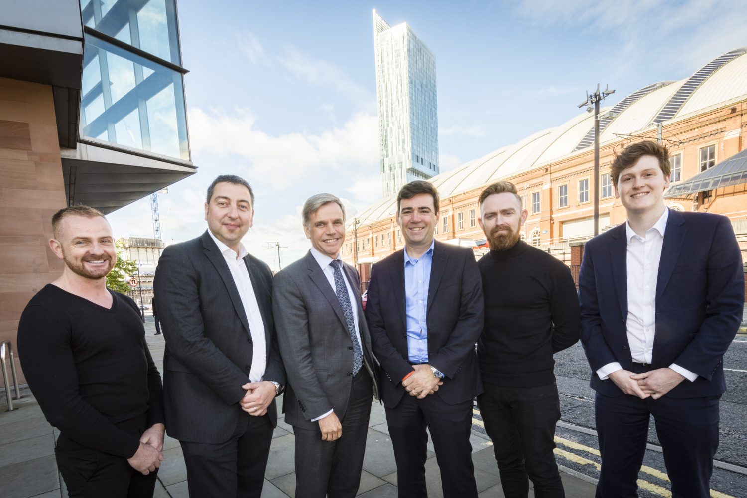 Six men standing outside an office building on a sunny day. The man to the far right is wearing a blue suit and white shirt, the man next to him is wearing black trousers and black jumper and has a ginger beard, the man to the far left has a brown beard and is wearing a V necked black jumper.