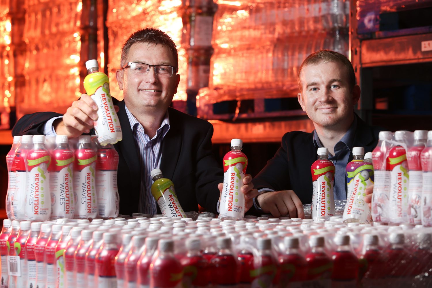 Two men in a factory surrounded by plastic bottles named Revolution. Man to the far left has glasses on and is wearing blue and white stripey shirt and is holding a plastic bottle.