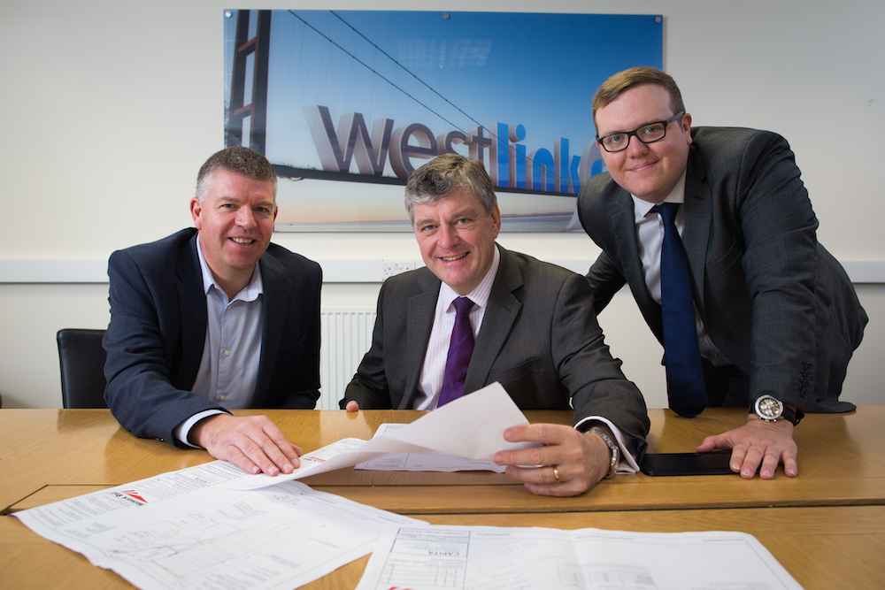 Three men in an office sitting behind a wooden desk. There is picture of their office branding directly behind them stating Westlinks. The man to the far right has his right hand on the table and is wearing glasses. He has a round face and is wearing a blue tie and a grey suit. The man in the middle has steel grey hair and is wearing a grey suit, pink suit and purple tie and the man to the far left is wearing a navy blue suit and light blue shirt.