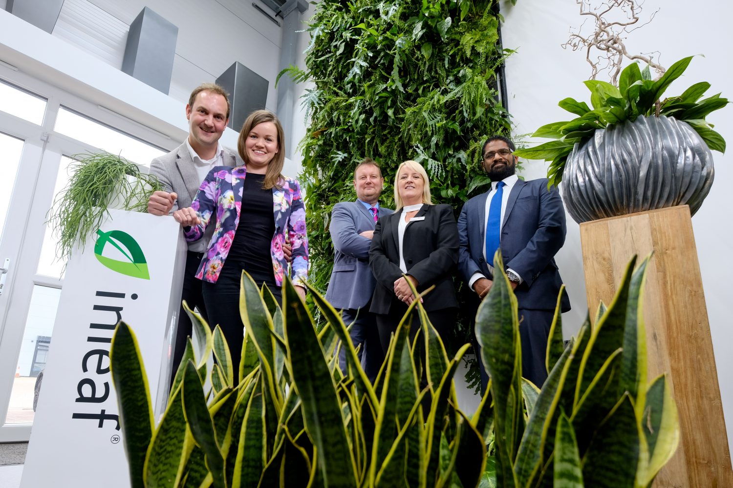 Five people standing in middle of foliage. Man at the left hand end standing next to company branding and to the right of him there is a lady wearing a colourful jacket and man at right hand end standing next to plant in a pot placed on wooden pillar.