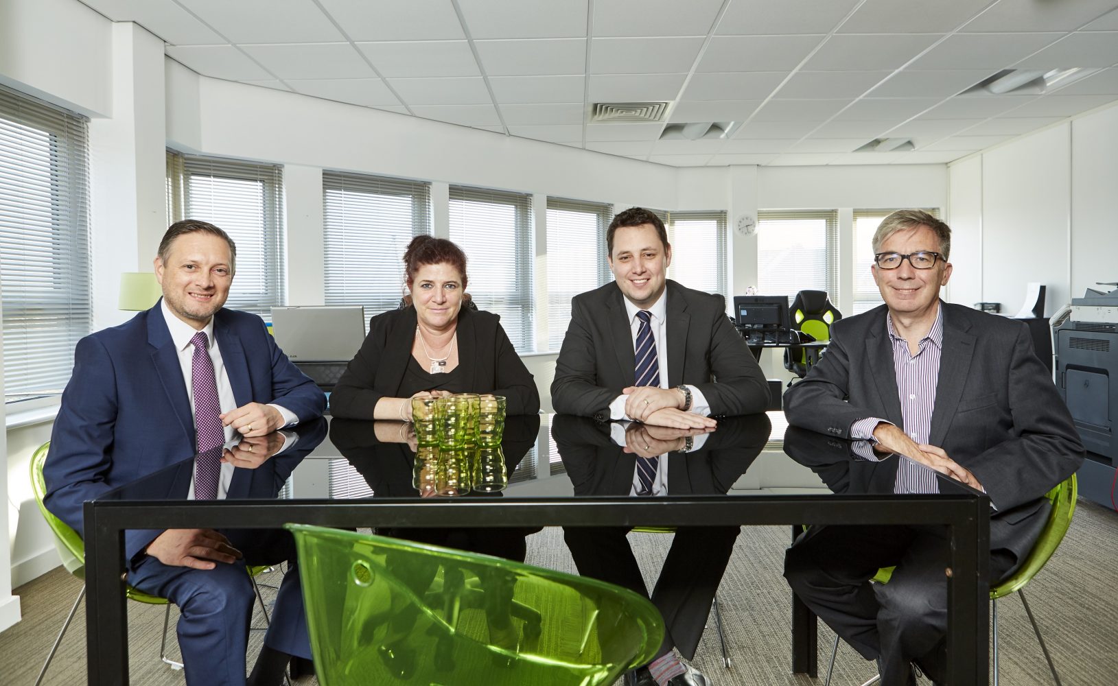 Three men and one woman wearing office sitting around an office table. They are sitting on green chairs.