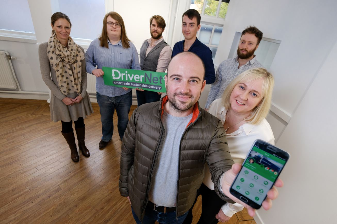 Two woman and five men wearing casual clothes in an empty office. Two people in the back row are holding Driver Net point of sale and two people at the front are holding a tablet.