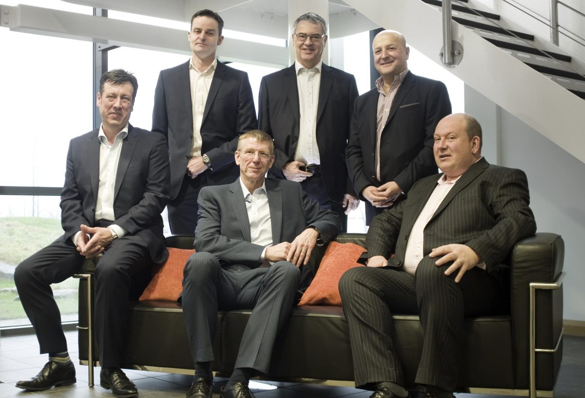 Group of six suited men in an office. Three of them perched on leather sofa and other three standing behind the sofa.