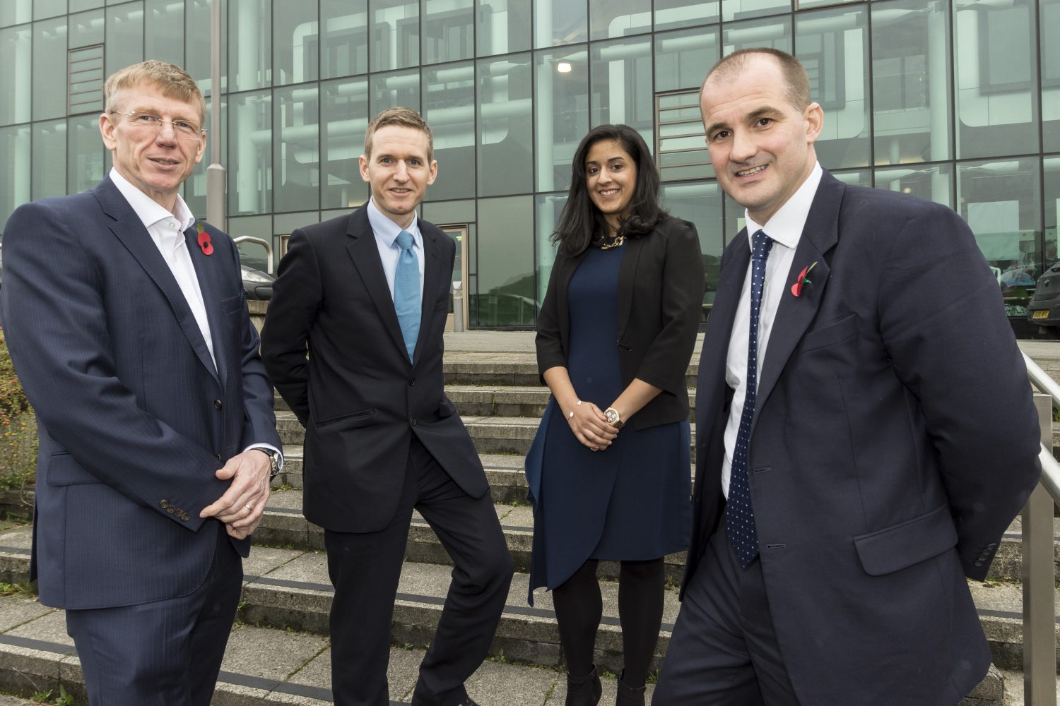 Four business executives standing on external steps in front of an office building. There is one woman in the group.