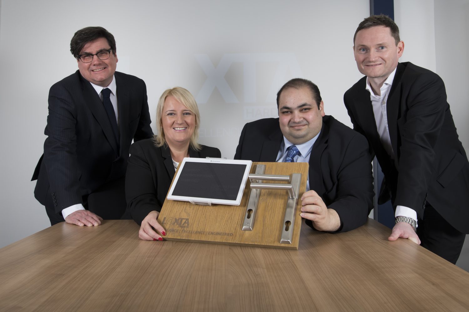 Four people around a desk. Man to the far right is wearing a black suit is standing, next to him is a seated man with his hand holding some equipment. There is a blond haired woman also holding the equipment. To the far left is man standing wearing glasses and a black tie.