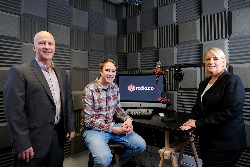 Two men and woman in black suit in a recording studio. Man in the middle of the group is wearing jeans and a casual shirt.