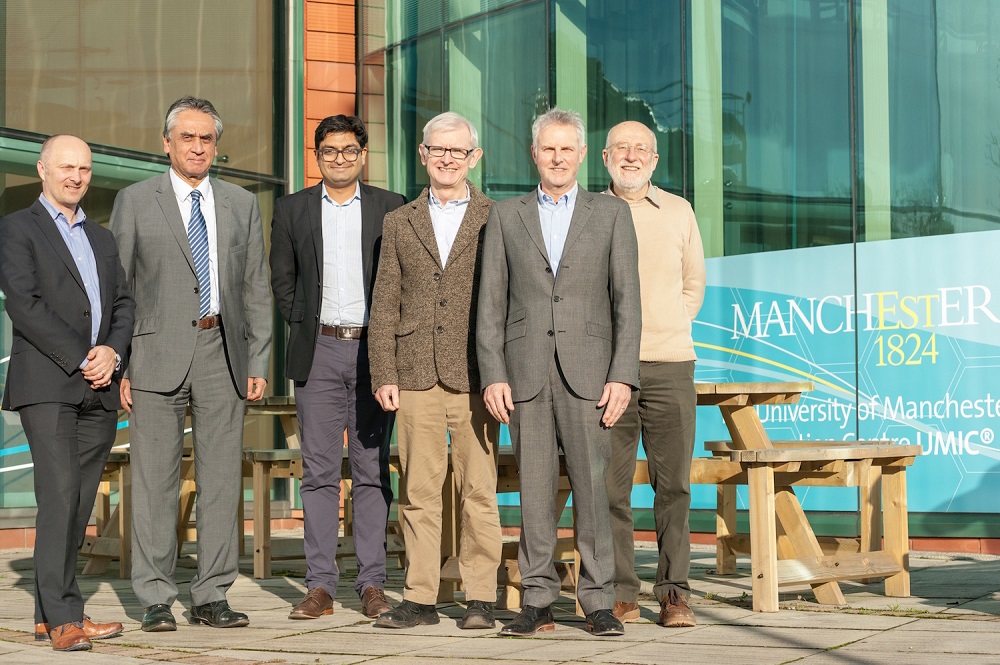 Six men in a row standing outside in front of a glass building. They are standing in front of pine tables and benches.