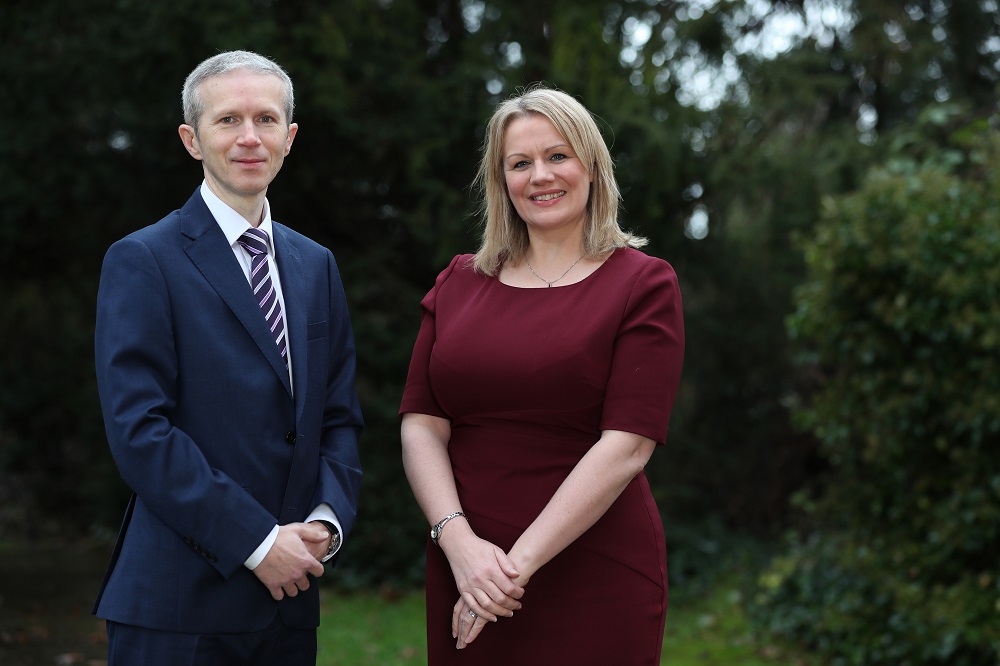 Businessman in garden setting standing next to business woman wearing a maroon dress.