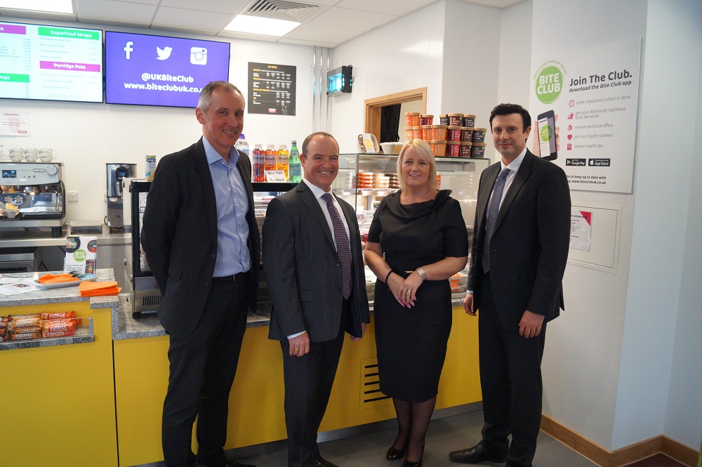 Three suited men and a business woman standing in front of office canteen food station.