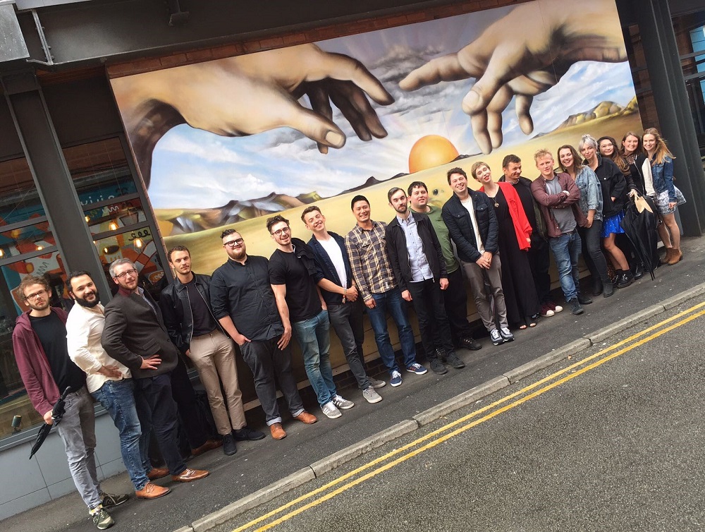 Large group of people standing in a row in front of poster of two large pair of hands and a sun in the middle. The people are standing on a pavement.