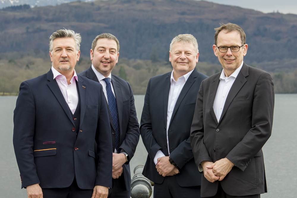 Four men outside with a view of mountains and a lake behind them. Man at the far right is wearing glasses, has a black suit and white shirt, man next to him is wearing a navy suit and has grey hair and the man at the far left has a pink shirt and is wearing a navy jacket.