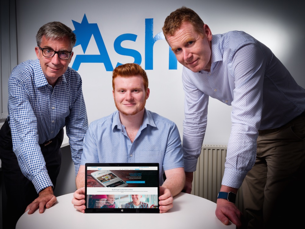 Three men in an office. Ginger haired man in the middle sitting down at a desk holding a tablet and two men either side of him both wearing blue and white checked shirts.