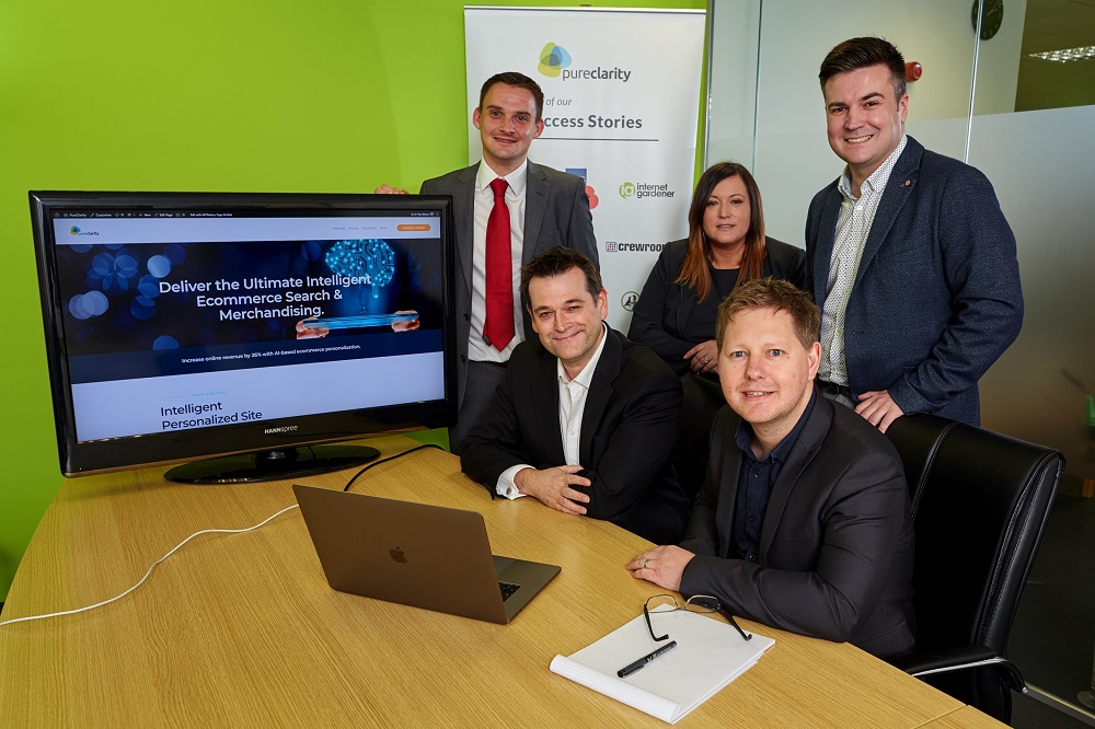 Five men and a woman in office. Two men are sitting at a desk. There is a large computer screen on the desk and the remaining three people are standing near the desk.