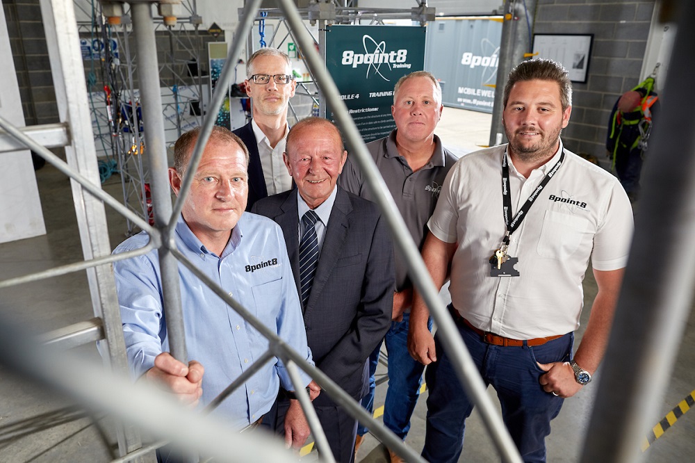 Five men in an office. Man to the far right is wearing a 8 Point 8 branded beige shirt, man in the middle of the group is wearing a navy suit and there is a man to the far left is holding onto a grey railing.