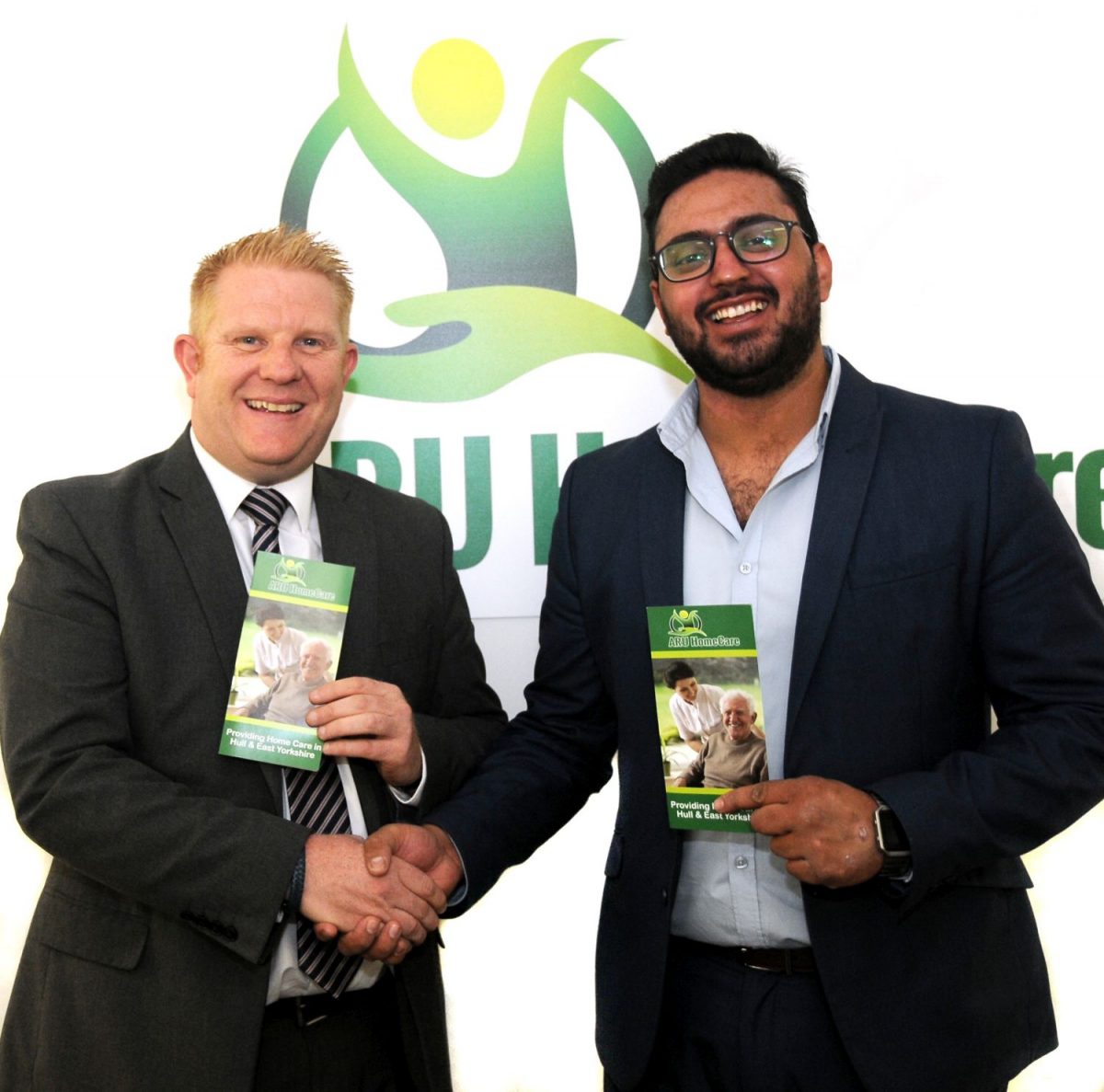 Two men holding leaflets shaking hands standing in front of Aru branding.