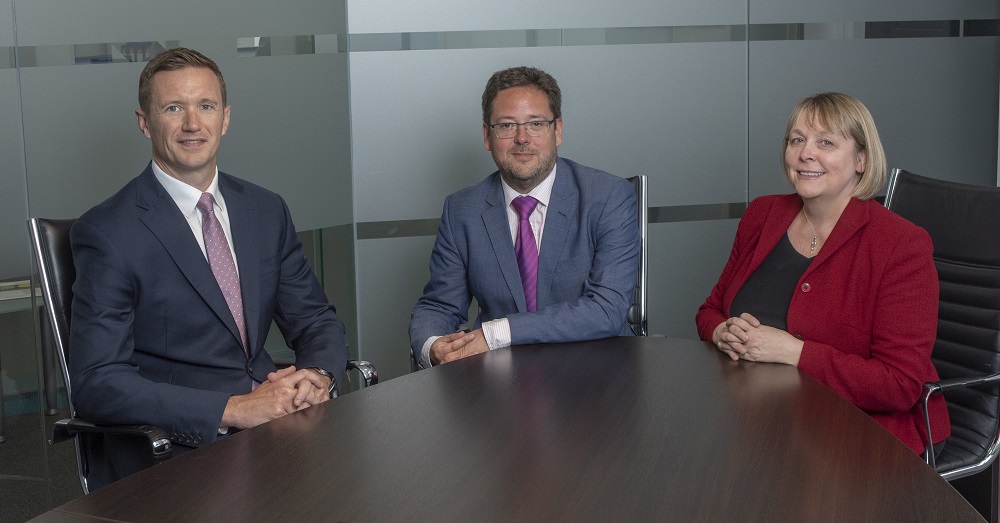 Three people in an office sitting around a desk. There is one woman in the group.