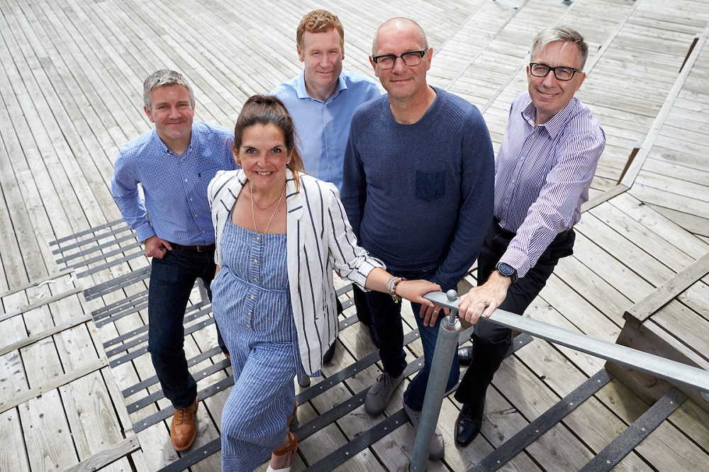 Five people standing outside. There is one woman who has her right hand on a hand rail. She is wearing a white and blue stripped jacket and a blue and white dress. The man directly behind her has grey hair and is wearing a blue shirt and there is a grey haired man wearing glasses with his hand on the handrail wearing a blue and white stripped shirt, black trousers