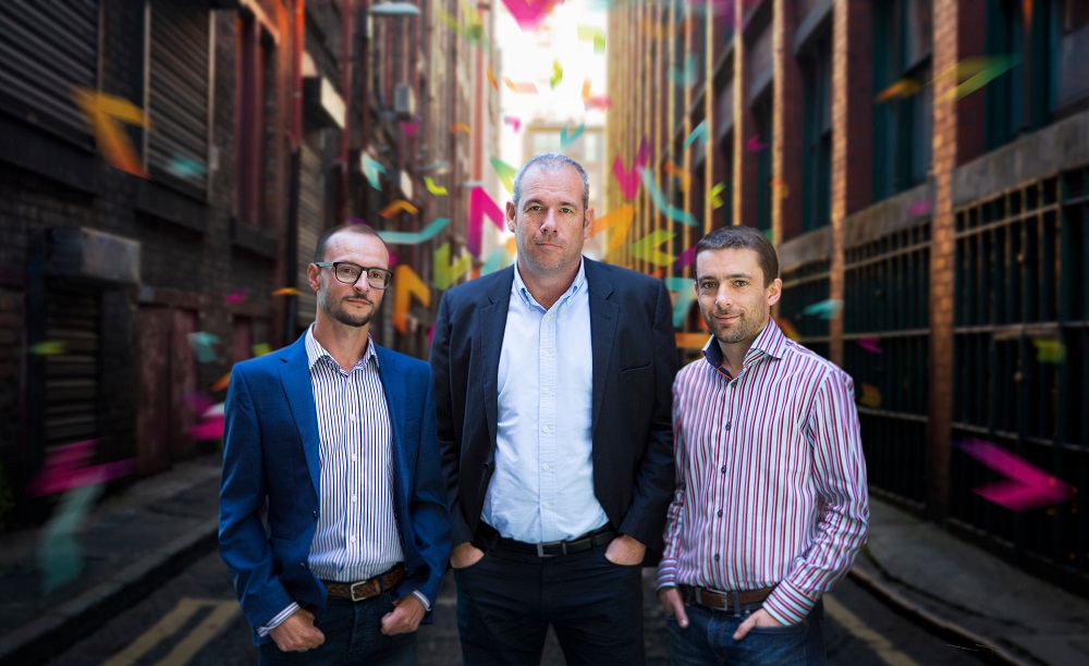 Three men outside in a street scene, they are sandwiched in the middle of tall office blocks. Man in the middle of the row is tall and is wearing a navy blue suit.