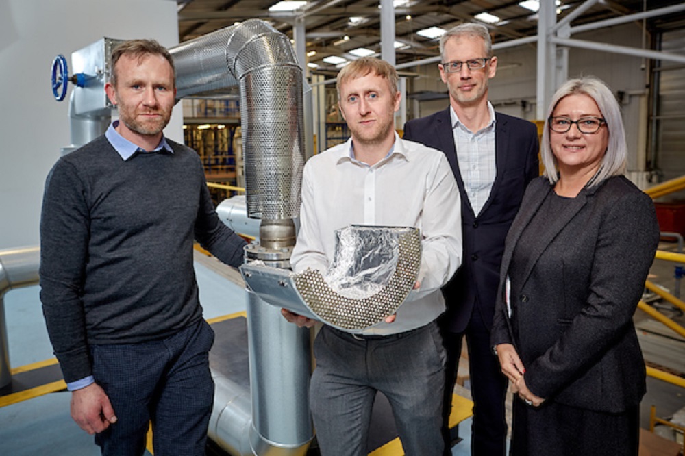 One woman wearing a black suit to the far right and three men in a factory. Man in the middle of the group is holding some industrial equipment.