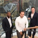 3 men stood on a staircase with a glass window in the background overlooking streets and buildings