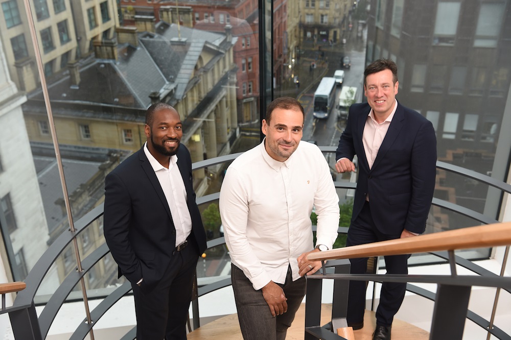 3 men stood on a staircase with a glass window in the background overlooking streets and buildings