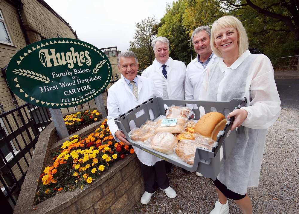 Three men wearing catering white clothing and one woman also wearing catering white clothing. One man and woman are holding a container full of bread.