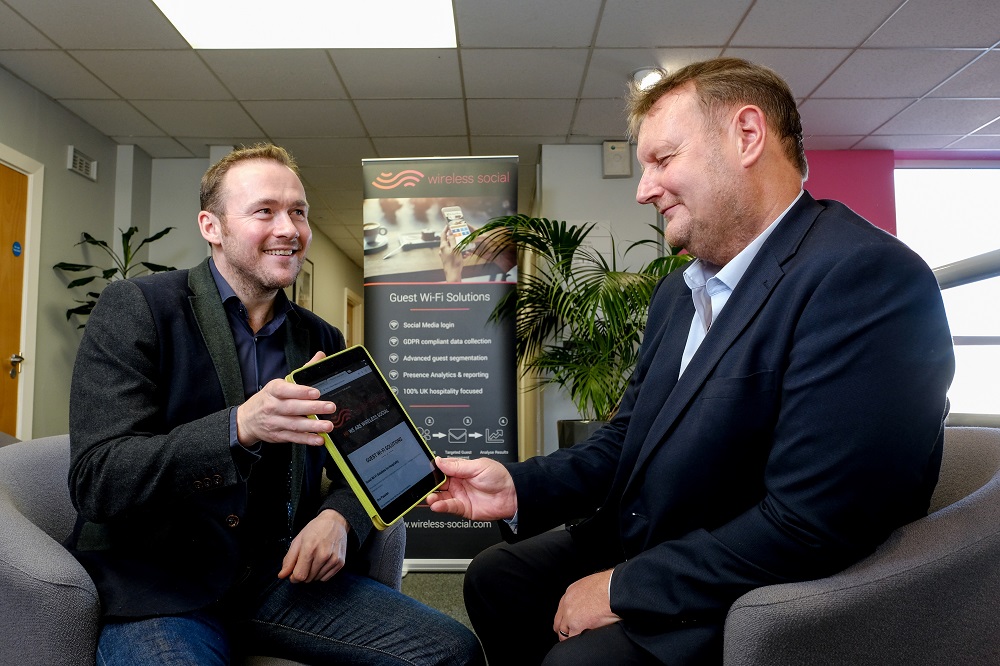 Two men in an office. Man to the right is wearing a blue suit and has his left hand holding a Tablet and the man opposite him is wearing a black jacket and blue jeans.