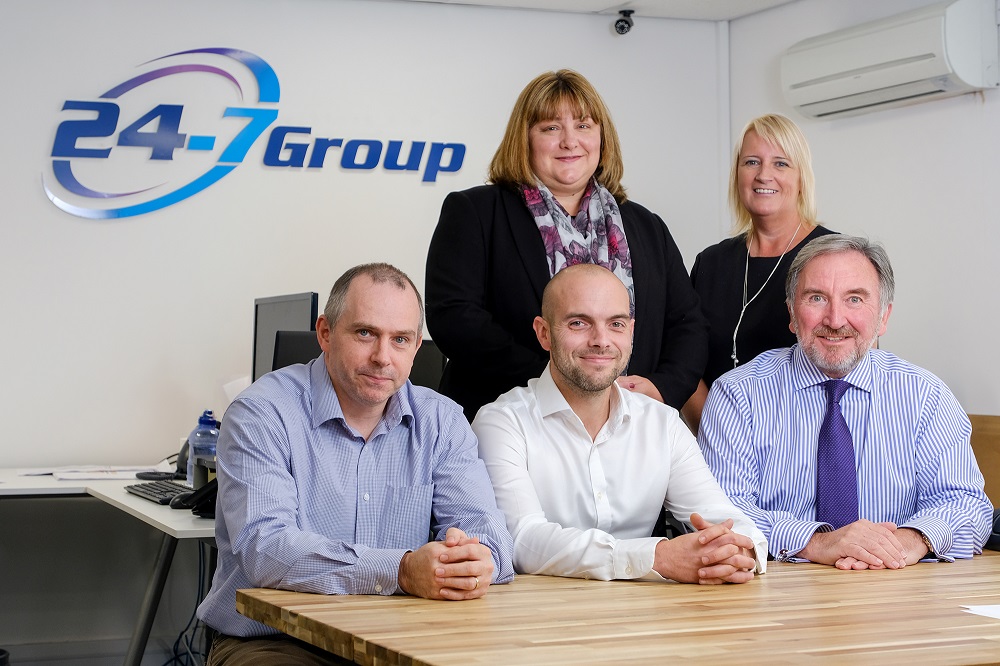 Five people in an office, three men sitting at a desk and two women standing up.