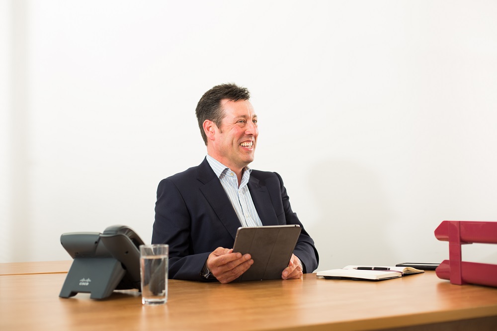 Will Clark, the Equity Fund Principal at Mercia sat a desk holding an iPad and laughing