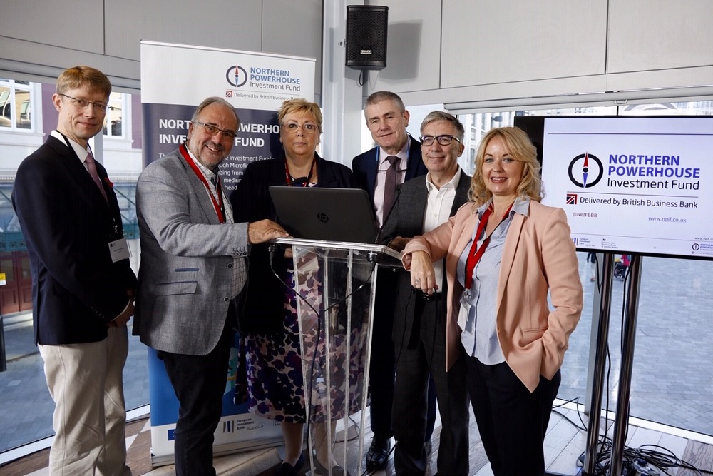 Four suited businessmen and two women in an office standing around a podium with Northern Powerhouse Investment Fund point of sale.