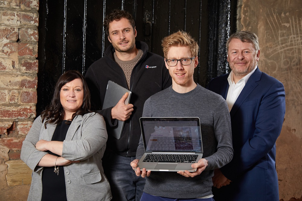 Four people standing in front of a black painted wooden wall and brickwork. Man to the far right has grey hair and is wearing a blue suit with a white shirt, man next to him is holding a laptop and there is one woman to the far left with long dark hair wearing a grey jacket.