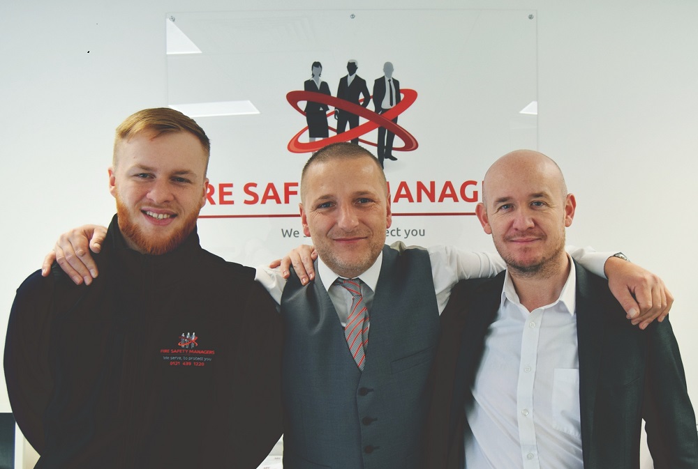 Three men standing together with arms around each others backs. They are standing in front of a poster.