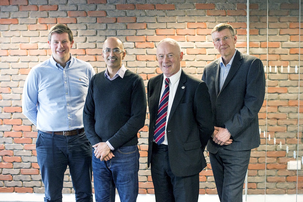 Four men standing in a row in an office in front of a brick wall.