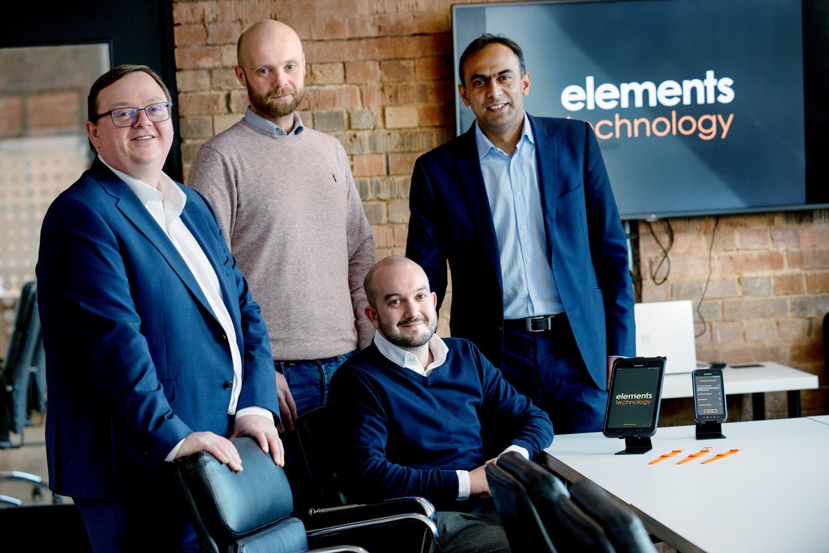 Four people in an office standing in front of a brick wall. There is one bearded man sitting in a chair next to desk and the other men are standing.