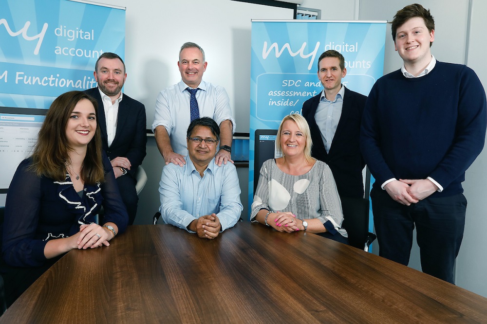 Seven people in an office standing and sitting around a table. Man to the far right is standing up wearing a black jumper, there are three people seated. The lady to the far left seated has long brown hair, next to her in the middle is an Asian man wearing a light blue shirt and to the right of him is a woman with mid length blond hair.