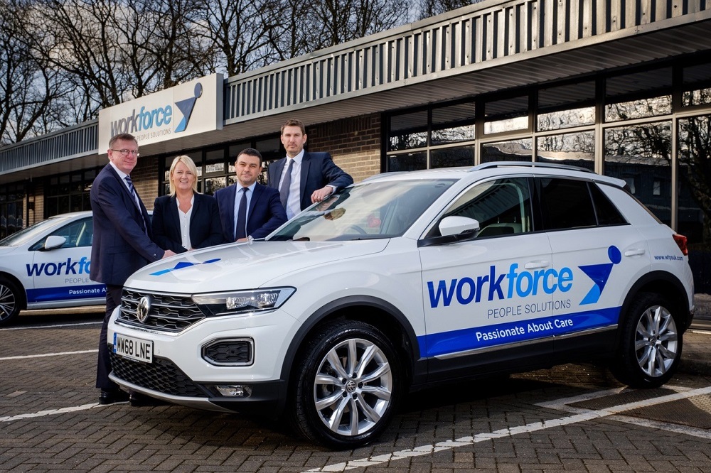 Four people standing behind a car. The car is branded Workforce People Solutions and it is Volkswagen SUV. There is one woman in the group. She has blonde hair and is wearing a black jacket and a white shirt and to the left of her is a man wearing a blue suit and glasses. At the far left there is a tall man wearing a blue suit and pale blue shirt.