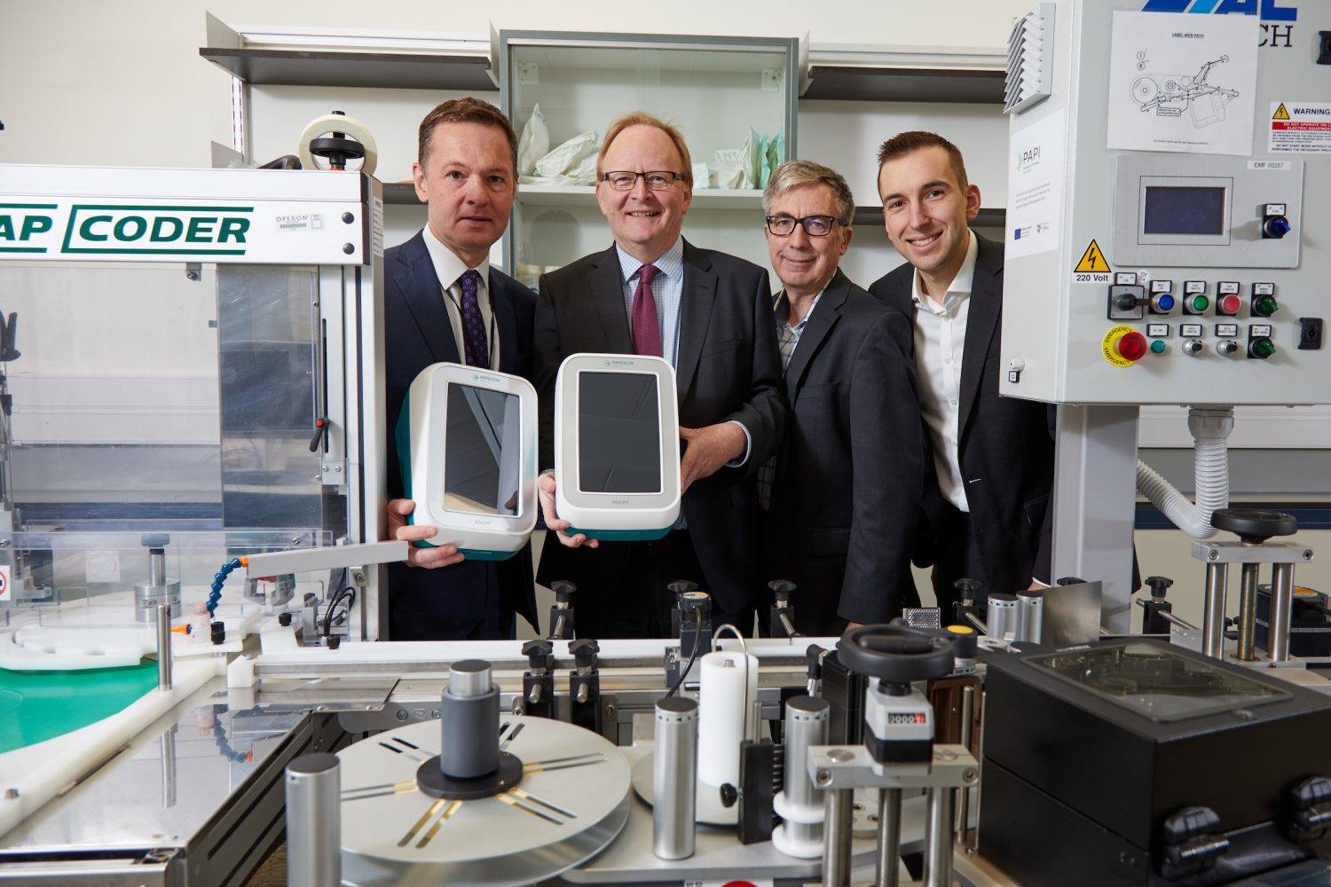 Four men in a laboratory in a row. Two men to the far left are holding equipment. Two men in the middle are wearing glasses. Young man to the far right has a black jacket and a white shirt.