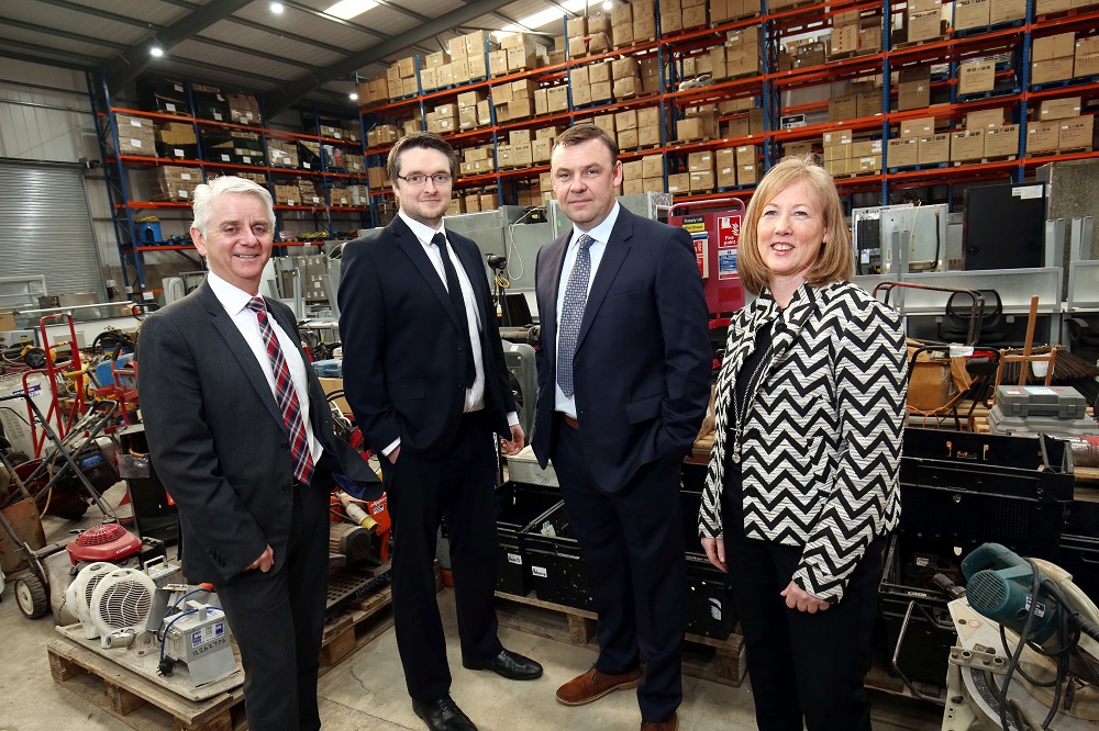 Woman in black and white jacket to the right and three men in suits. They are in a factory.