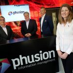 Three men wearing black suits and a woman wearing white shirt and black trousers standing in a room with Phusion Information Management point of sale.