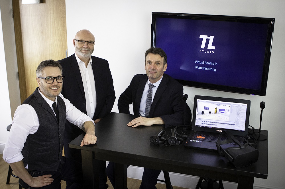 Two men sitting in an office and one bearded man with black suit standing. They are in an office with TL Studio on a TV screen behind them.