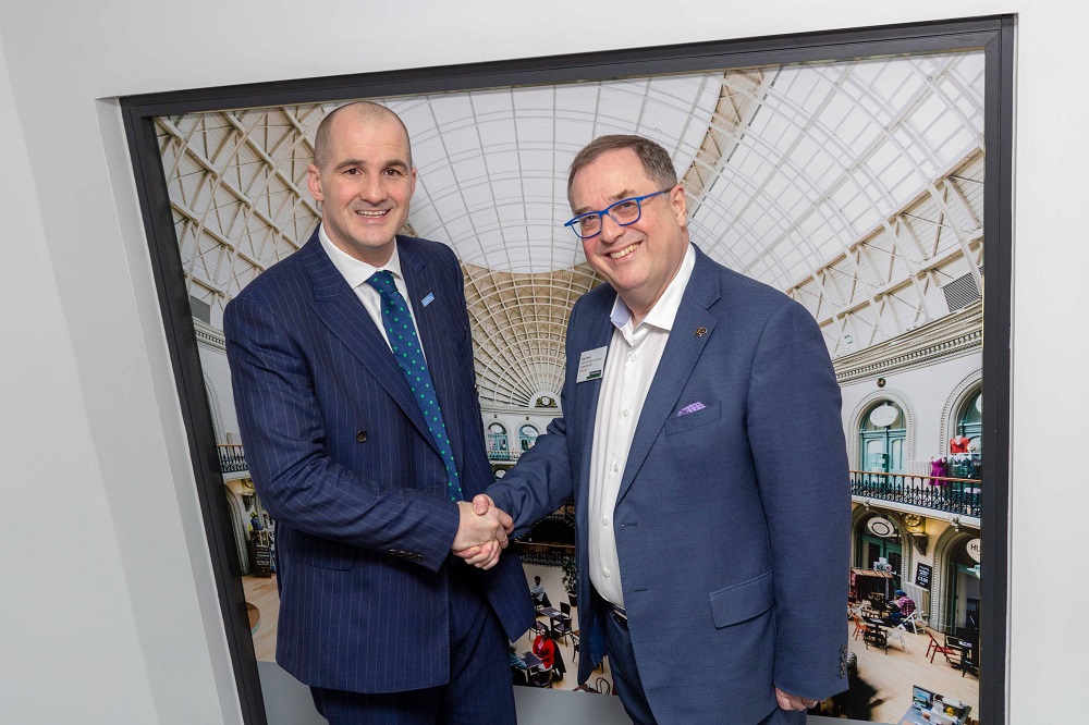 Two men shaking hands. Man to the far right has blue rimmed glasses and is wearing a blue suit and the other man is wearing a blue spotty tie.