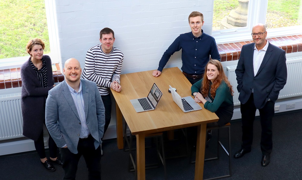 Six people in an office, two men and a woman one side of a desk and two men and a woman other side of desk.