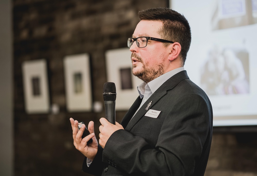 Man with dark hair a glasses holding a microphone standing in front of brick wall with pictures.